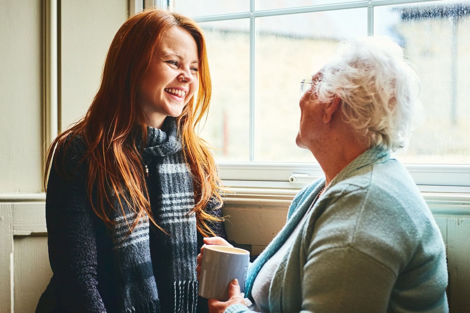 A young woman laughing with an older woman who holds a mug of tea, by a window